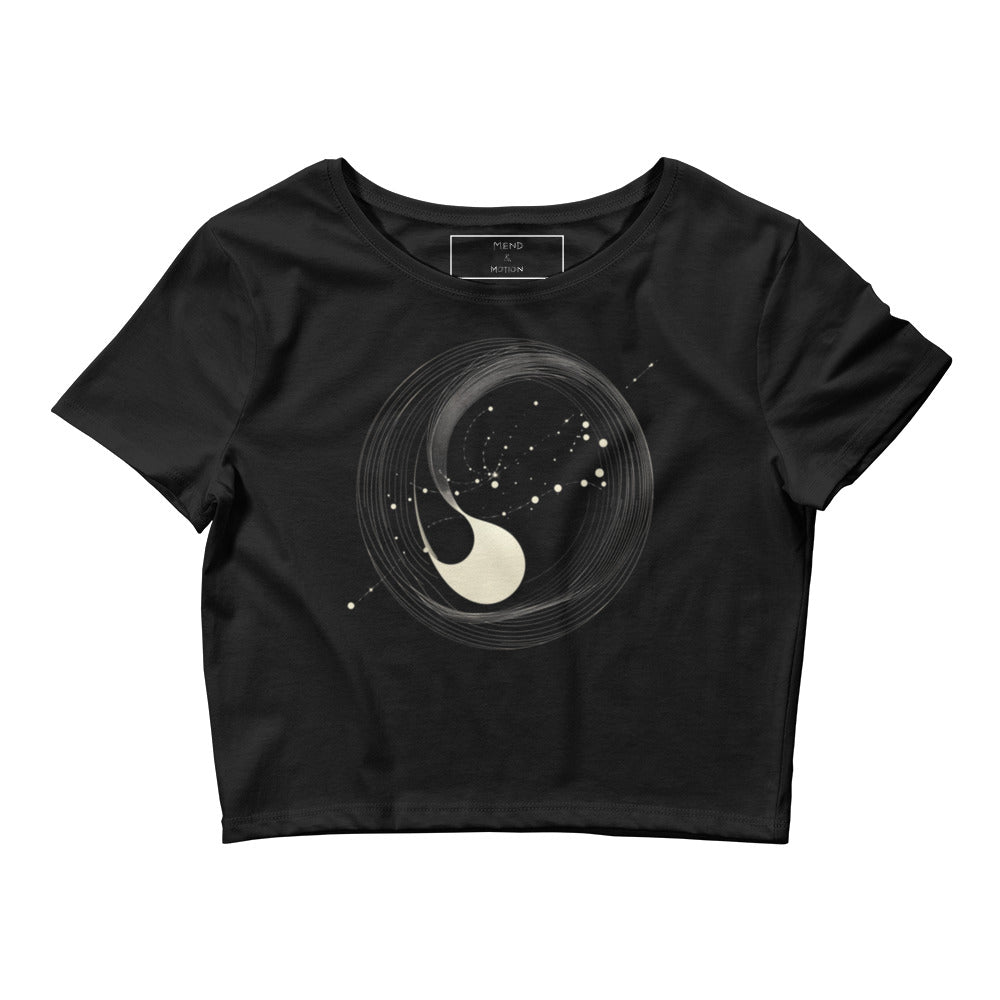 A flat lay of a black women's crop tee featuring an abstract yin-yang galactic design with a swirling celestial theme.