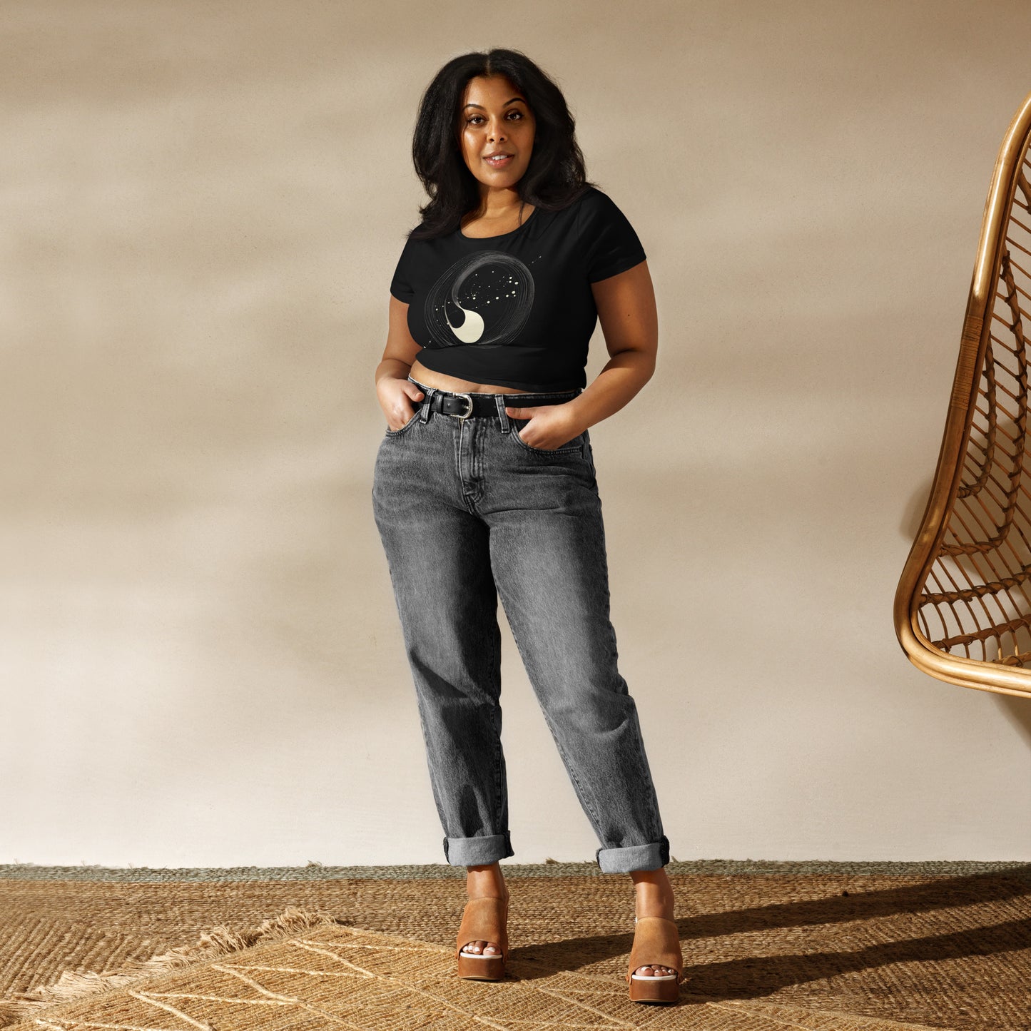 plus-size woman wearing a black crop tee with an abstract yin-yang celestial design, styled with gray jeans and heels in a neutral indoor setting.