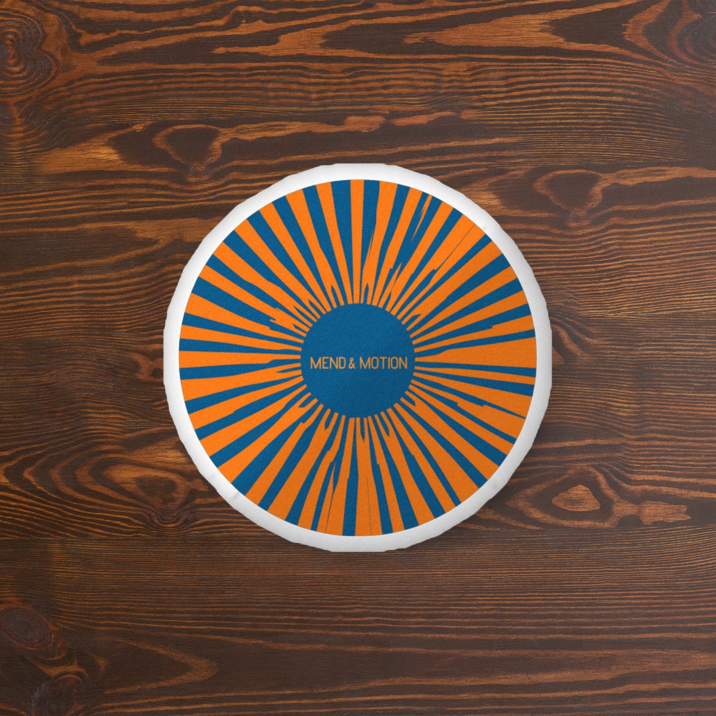Circular pillow with Mend & Motion logo and orange-blue radial design on a wooden background