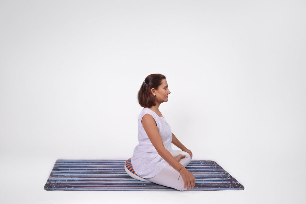A woman practicing meditation in a seated position on a Mend & Motion cushion, placed on a striped yoga mat against a clean white background.