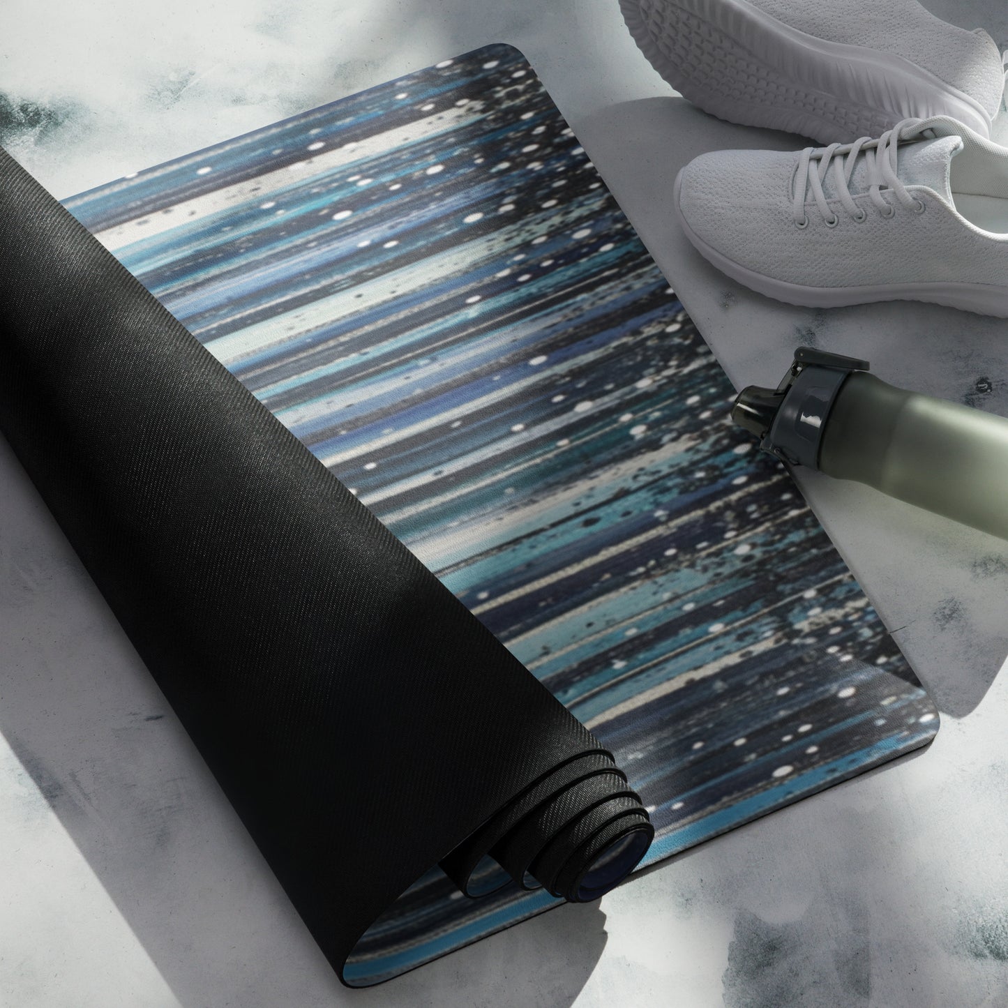 A rolled yoga mat with a serene blue and black streaked design, accompanied by white shoes and a water bottle on a light surface
