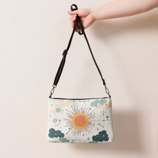 Hand holding a Mend & Motion crossbody bag featuring a celestial sun, clouds, and starry pattern on a cream background.