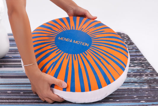 A round meditation cushion with an orange and blue sunburst design and the "Mend & Motion" logo, placed on a yoga mat.